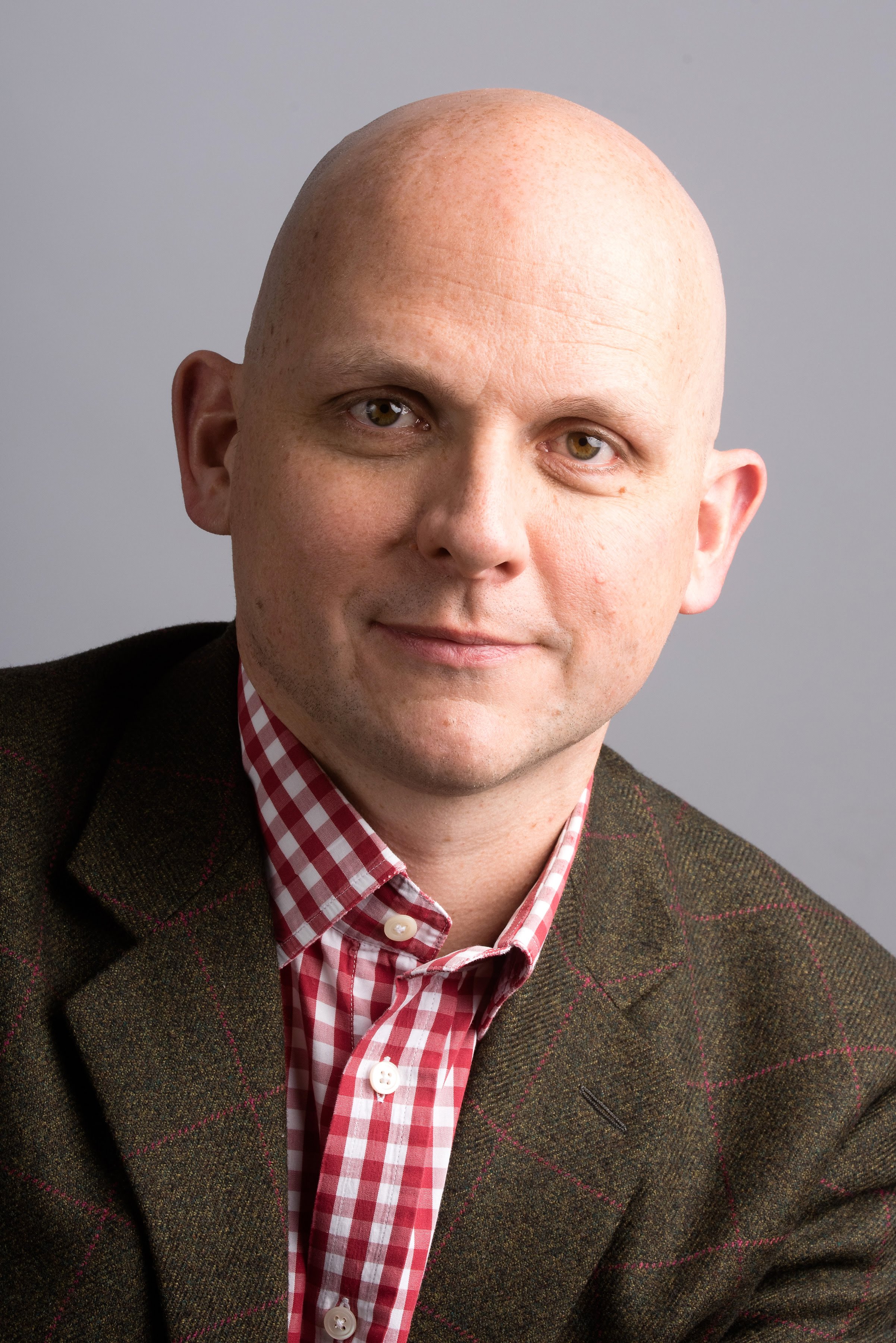 Image of Professor Gunter Kuhnle in front of a grey background wearing a green tweed jacket over a red and white checked shirt. 