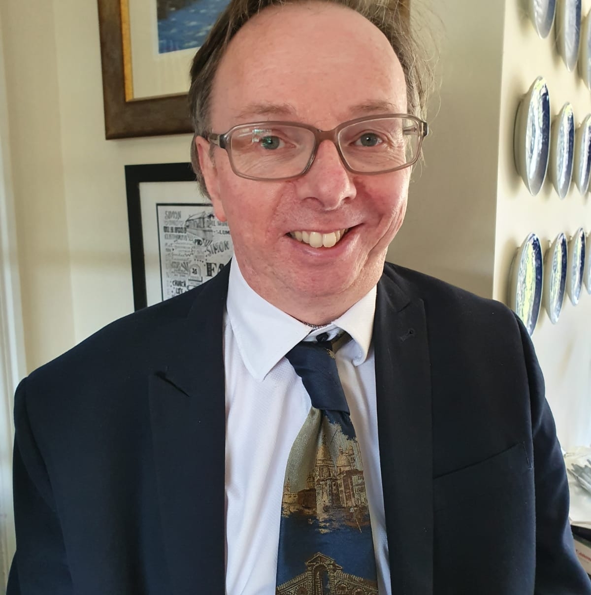 Image of Dr Simon Wilkinson in front of a cream coloured wall with decorations, wearing a dark suit jacket over a white shirt and multicoloured tie. 