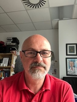 Photo of Professor Gareth Jenkins- Chair.  The photo is a head and shoulders image. Gareth is wearing a red shirt, dark framed glasses  and has facial hair. In the background the image shows a ceiling covered in white square tiles, to the left a book shelf and to the right, framed pictures on the wall. 