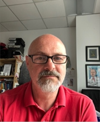 Professor Gareth Jenkins. Gareth is wearing a red shirt, has grey facial hair and is wearing dark rimmed glasses. Gareth is photographed in an office with wall art and shelving behind him. There are square ceiling tiles above him.