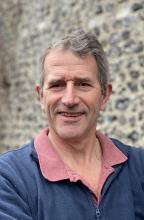 Image of Lay Member Nick Richardson. Nick is photographed in front of a blurred  stone wall. Nick has  short greyish brown hair and is wearing a navy blue zip up jumper and a dark pink collared shirt.    